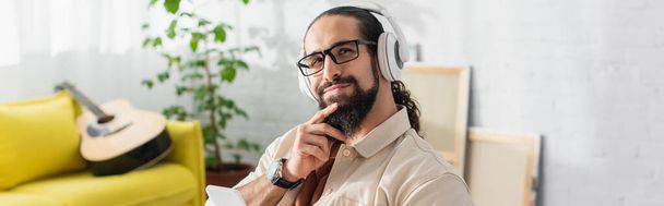 мрійливий іспаномовний чоловік в навушниках посміхається, слухаючи музику біля гітари на розмитому фоні, банер
 - Фото, зображення