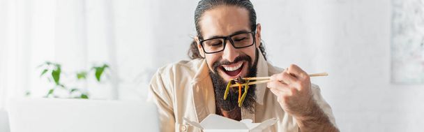freelancer hispano positivo en anteojos comiendo fideos chinos cerca de la computadora portátil borrosa, pancarta - Foto, Imagen