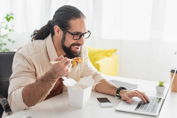 χαμογελαστός ισπανόφωνος ελεύθερος επαγγελματίας δακτυλογραφεί στο laptop ενώ τρώει κινέζικα noodles στο σπίτι - Φωτογραφία, εικόνα