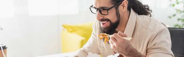 hispanic man in eyeglasses smiling while eating chinese noodles at home, banner - Photo, Image