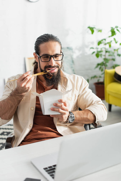 freelancer latino barbudo olhando para laptop enquanto come macarrão chinês para o almoço em casa - Foto, Imagem