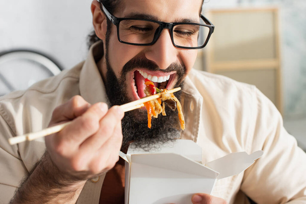 bearded latin man in eyeglasses eating chinese noodles with sticks on blurred foreground - Photo, Image