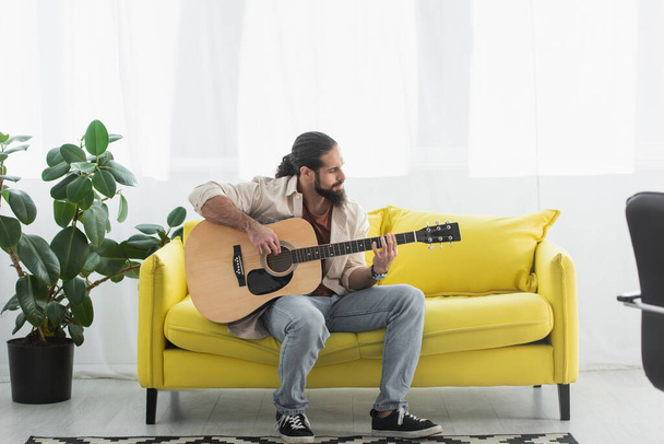 full length view of latino man sitting on yellow couch in living room and playing guitar - Foto, Imagem