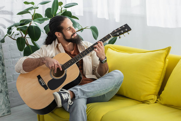 латинос играет на гитаре, сидя дома на жёлтом диване - Фото, изображение