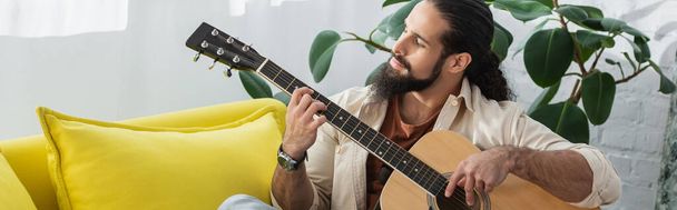 bebaarde Spaanse man die thuis op de bank zit en akoestische gitaar speelt, banner - Foto, afbeelding