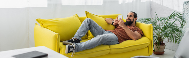 Full length άποψη της Λατινικής άνθρωπος ξαπλωμένος σε κίτρινο καναπέ και κουβέντα στο smartphone, πανό - Φωτογραφία, εικόνα