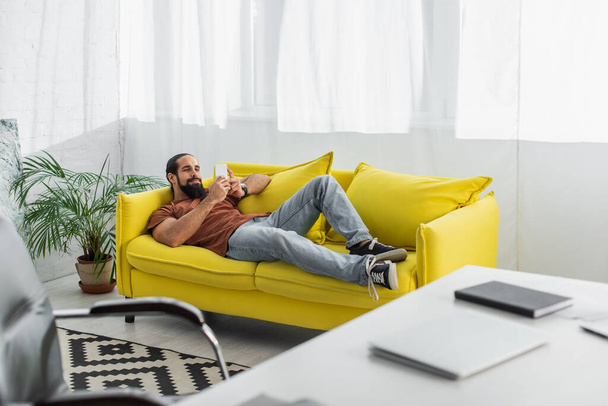 Ισπανόφωνος ελεύθερος επαγγελματίας κουβεντιάζοντας στο smartphone, ενώ στηρίζεται σε καναπέ κοντά θολή γραφείο με φορητό υπολογιστή - Φωτογραφία, εικόνα