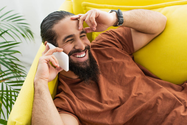 χαρούμενος ισπανόφωνος άνθρωπος αγγίζοντας μέτωπο, ενώ μιλάμε στο κινητό τηλέφωνο στο σπίτι - Φωτογραφία, εικόνα