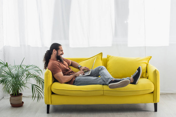 side view of hispanic man sitting on yellow couch at home and using laptop - Foto, afbeelding