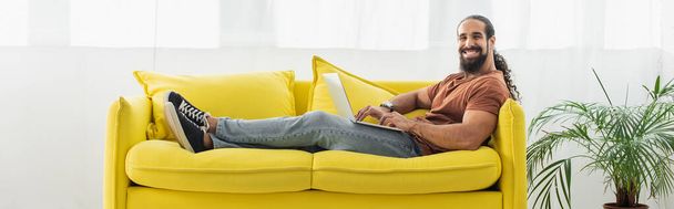 веселый латиноамериканец сидит на желтом диване с ноутбуком и улыбается в камеру, баннер - Фото, изображение