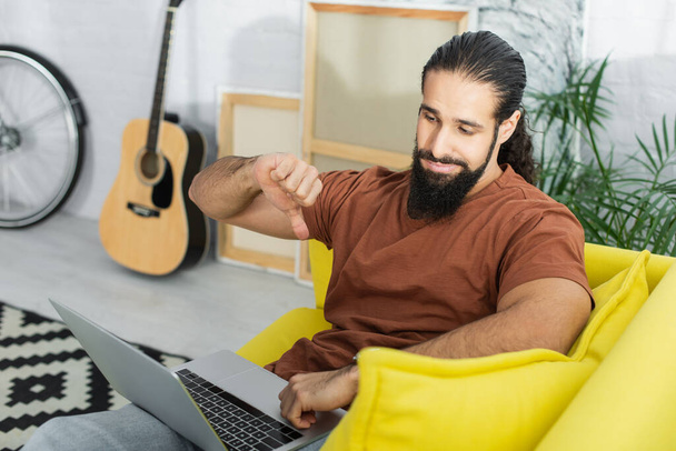 Ισπανόφωνος άνθρωπος δείχνει απέχθεια χειρονομία, ενώ χρησιμοποιείτε φορητό υπολογιστή στον καναπέ κοντά θολή κιθάρα και καμβάδες - Φωτογραφία, εικόνα