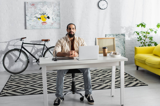 bärtiger hispanischer Freiberufler sitzt am Arbeitsplatz im Heimstudio - Foto, Bild