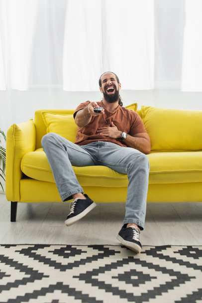 ενθουσιασμένοι Λάτιν άνθρωπος με τηλεχειριστήριο γέλιο σε κίτρινο καναπέ, ενώ βλέποντας τηλεόραση - Φωτογραφία, εικόνα