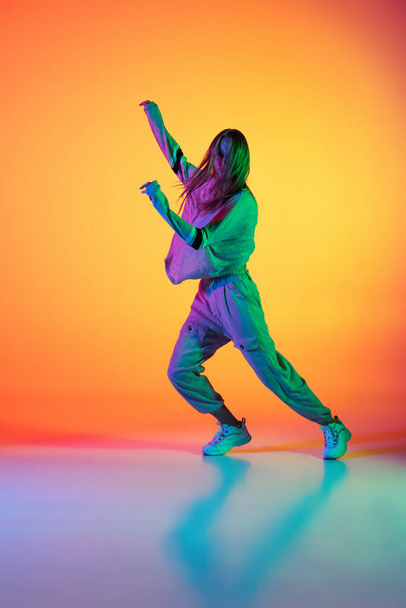 One Caucasian sportive girl dancing hip-hop in stylish clothes on colorful background at dance hall in neon light. Youth culture, movement, style and fashion, action. - Photo, Image