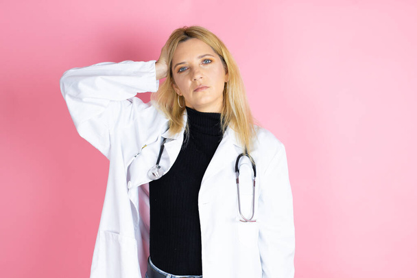 Young blonde doctor woman wearing stethoscope standing over isolated pink background putting one hand on her head smiling like she had forgotten something - Photo, Image