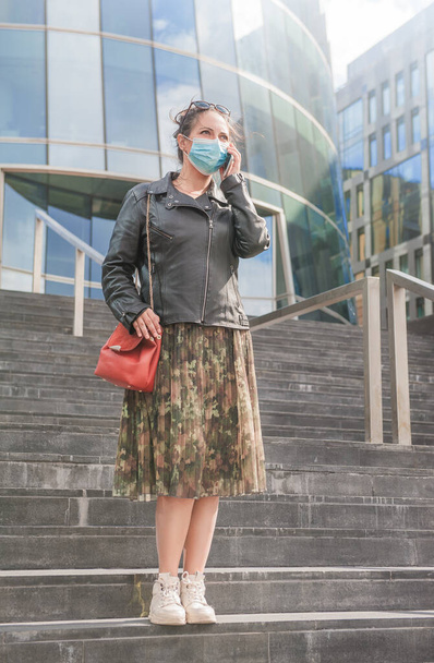 Mulher bonita vestindo máscara médica falando no telefone móvel de pé perto da parede do espelho de fundo do edifício da cidade - Foto, Imagem