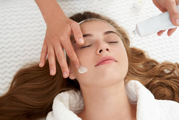 Cosmetologist applying cream on female face. Woman having a facial treatment in spa salon - Fotó, kép
