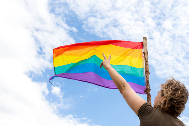 Lockige blonde Frau, von der Seite nicht wiederzuerkennen, die eine Regenbogenfahne schwenkt. Zwei Finger im Siegeszeichen auf blauem Himmelshintergrund mit Wolken. LGBTQA-Konzept - Foto, Bild