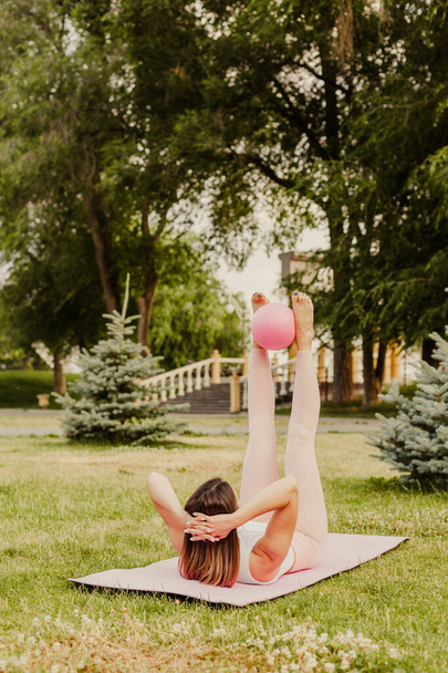 athletic, slender woman in pink clothes and small rubber ball doing Pilates on pink mat at dawn in park in summer - 写真・画像