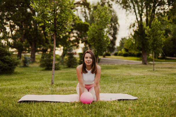 Charming fit young woman is sitting on knees on sports mat with rubber small pilates ball in park - Photo, image