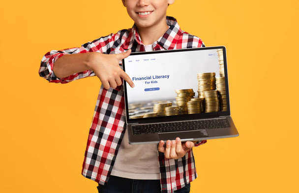 Close-up van gelukkige jongen wijzen naar laptop scherm met financiële geletterdheid voor kinderen educatieve website, oranje achtergrond - Foto, afbeelding
