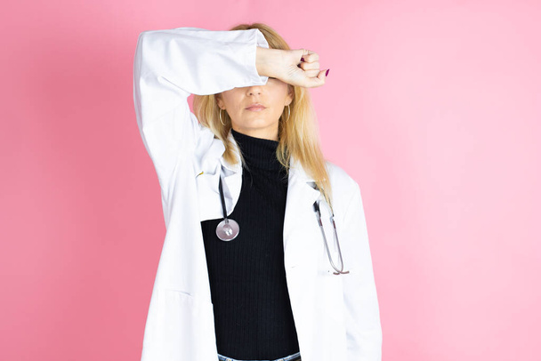 孤立したピンクの背景に立って聴診器を身に着けている若いブロンドの医師の女性は陽気で面白い笑顔で目をカバーしています.ブラインドコンセプト. - 写真・画像