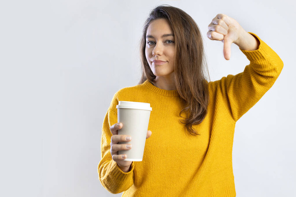 Young woman with wearing yellow sweater holding a cup of coffee with angry face, negative sign showing dislike with thumbs down, rejection concept - Foto, Bild