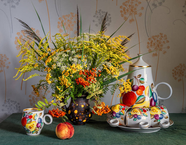 Um buquê de flores de prado e ramos de Rowan com cachos vermelhos de bagas. Na mesa está um jarro grande, copos e um pêssego maduro. - Foto, Imagem