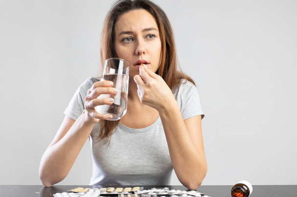 Young sick woman taking pills holding a glass of water on gray background - Fotoğraf, Görsel
