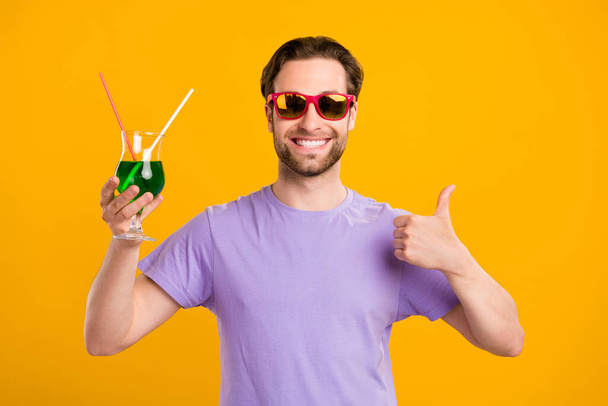 Photo of attractive cute gentleman wear violet t-shirt dark eyewear drinking cocktail showing thumb up isolated yellow color background - 写真・画像