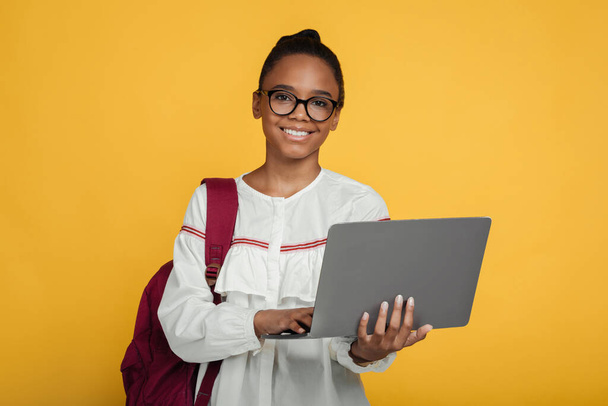 Smiling smart cute teenage african american girl pupil in glasses with backpack studying with laptop - Foto, Bild