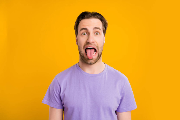 Photo of childish fooling young guy dressed purple t-shirt showing long tongue isolated yellow color background - Foto, immagini