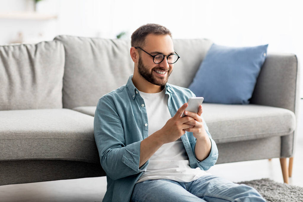 Chico joven feliz en gafas usando el teléfono celular, sentado en el suelo en la sala de estar, trabajando o comunicándose en línea desde casa - Foto, imagen