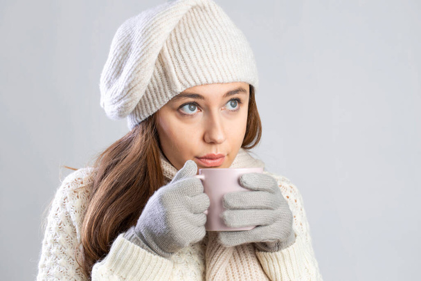 Young woman in knitted sweater and beanie hat with a mug in hands at grey background. Warming up concept. - Foto, immagini