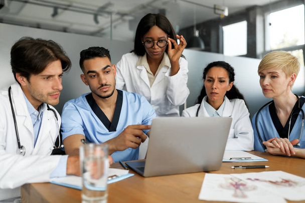 Skoncentrowany wielonarodowy zespół lekarzy siedzących przy stole, korzystających z laptopa - Zdjęcie, obraz