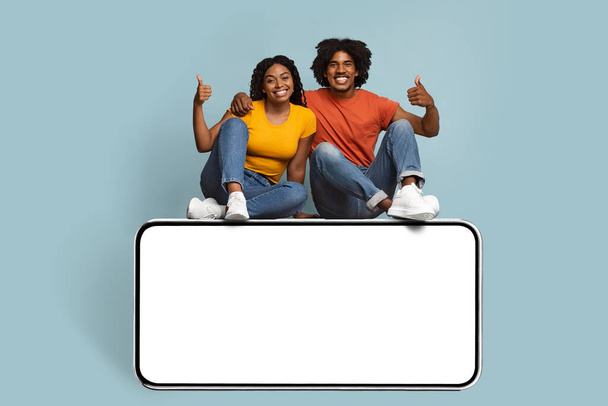 Positive black couple sitting on big smartphone with blank screen - 写真・画像
