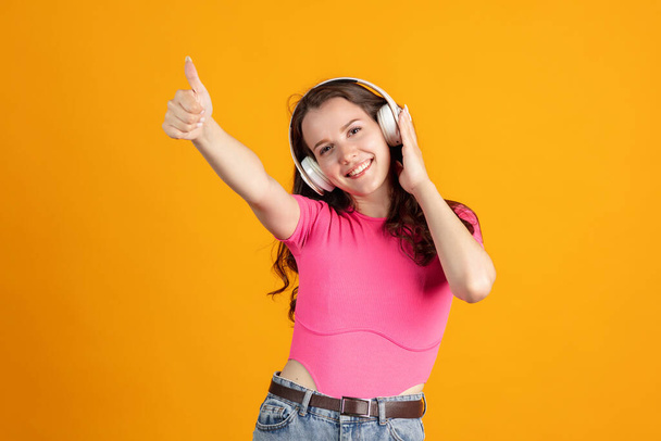 Cropped portrait of young beautiful girl listening to music in headphones with thumbs up isolated over orange studio background - Photo, image