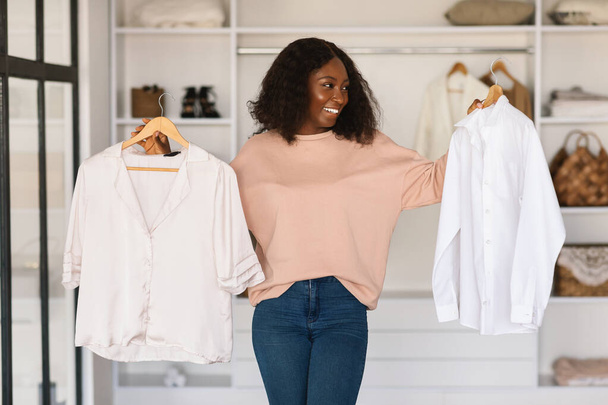 Happy African Woman Shopping And Choosing Clothes Holding Shirts Indoors - Foto, Imagen