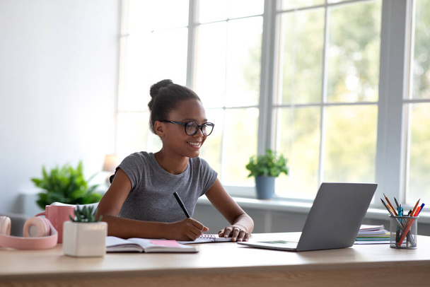 Sorridente adolescente studente ragazza nera in occhiali studiare a casa con il computer portatile e guardare lezione online - Foto, immagini