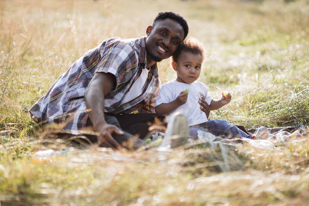Happy african man sitting in hugs with little son outdoors - Фото, зображення