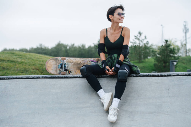 Joyful woman skater with short hair in sunglasses resting on a deck, enjoying herself. She wears some protective gear. Frontal view. Nature in background. - Fotó, kép