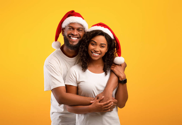 Vrolijke zwarte echtgenoten in Santa hoeden knuffelen op gele studio achtergrond. opgewonden Afrikaans amerikaanse man omarmen zijn mooie vrouw van achteren, vieren Nieuwjaar of Kerstmis samen - Foto, afbeelding