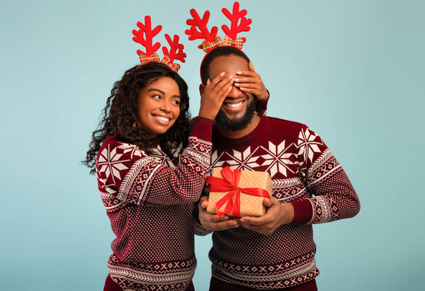 Loving black wife making New Year gift for her husband, covering his eyes. African american couple wearing winter sweaters and deer horns, celebrating Christmas, blue studio background - Foto, immagini