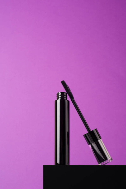 Open zwarte mascara tube met een transparante hoes en penseel op een zwart podium en paarse achtergrond. Make-up product voor ogen met ruimte voor tekst. - Foto, afbeelding