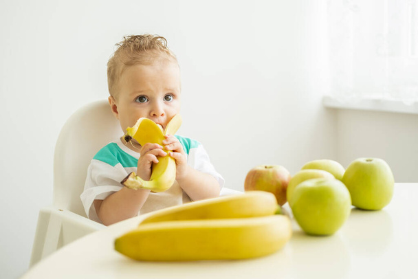 子供の椅子に座って白いキッチンでバナナを食べる面白い赤ちゃん - 写真・画像