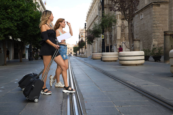 Dwie młode i piękne kobiety zwiedzające miasto. Kobiety przemierzają dużą aleję na tramwaju. Mają walizki na kółkach, mapy i kamery. Pojęcie urlopu i podróży. - Zdjęcie, obraz