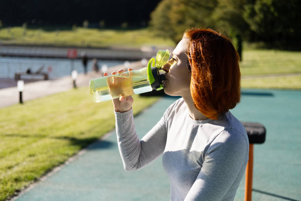 mujer de jengibre en ropa deportiva beber agua mientras se trabaja en el campo de deportes en el día soleado otoño. - Foto, imagen