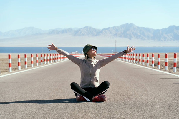 asian woman tourist sitting in the middle of an empty open road with lake and rolling mountains in background, leg crossed arms outstretched. - Photo, Image