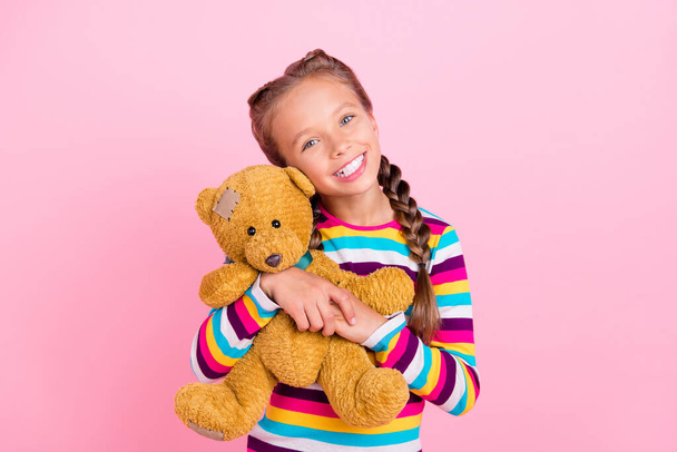 Photo of cheerful dreamy small lady hug toy bear shiny smile wear striped shirt isolated pink color background - 写真・画像