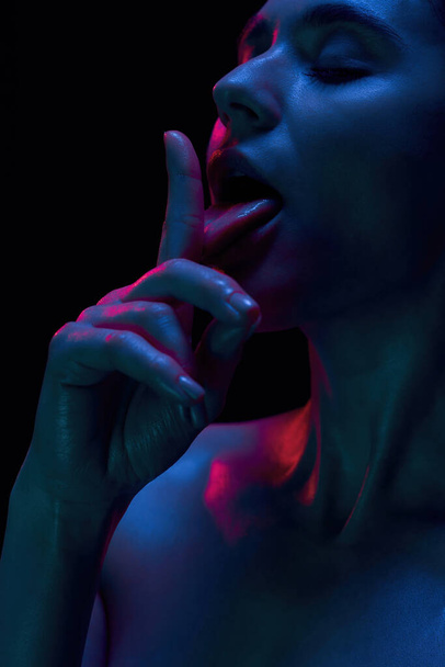 Closeup of provocative naked young woman licking her finger, posing isolated in blue pink neon light over black background - Φωτογραφία, εικόνα